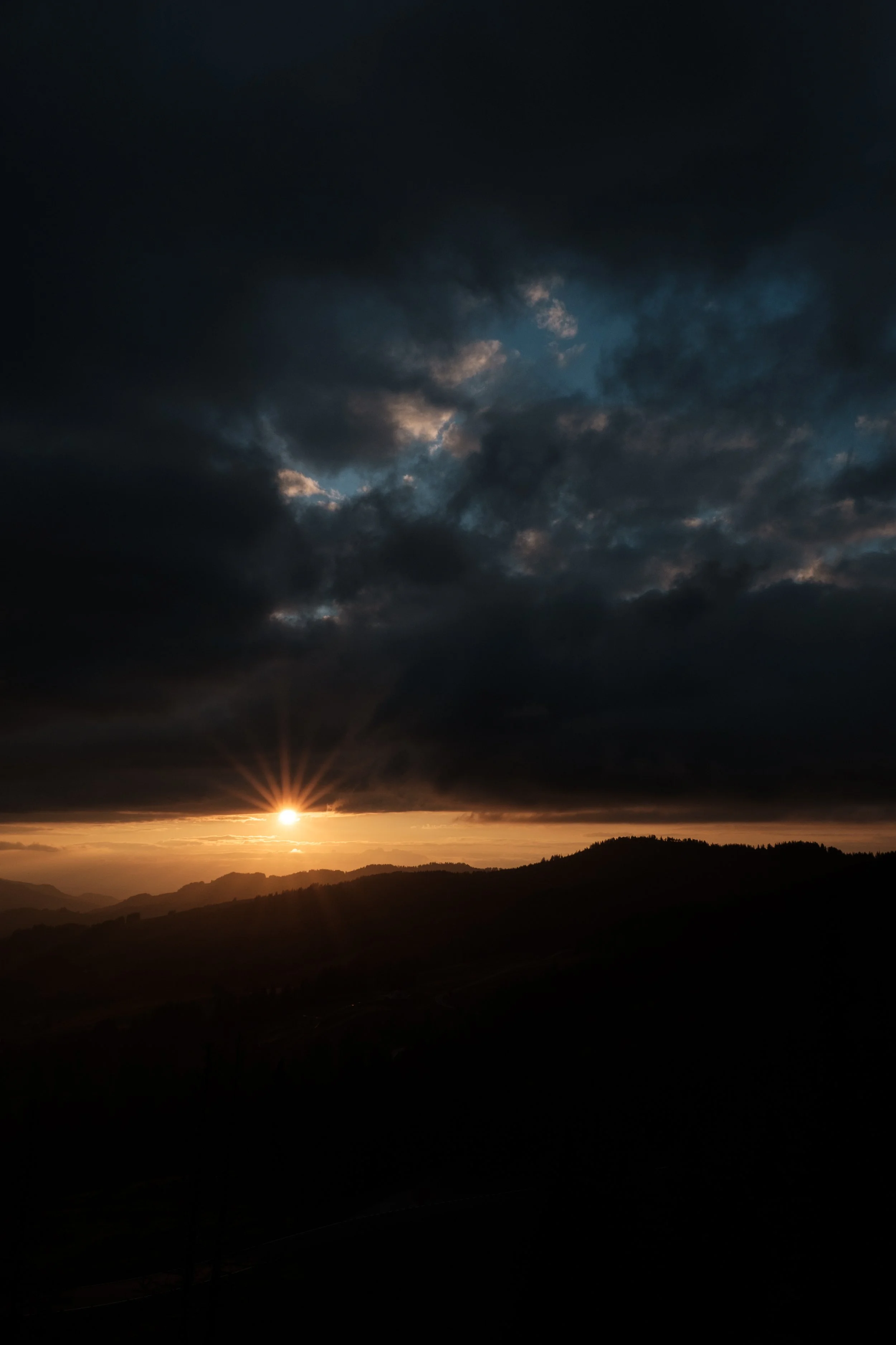 Sonnenuntergang hinter Hügeln, dunkle Wolken am Himmel, Sonnenstrahlen brechen durch die Wolken, Landschaft mit Hügeln im Vordergrund.