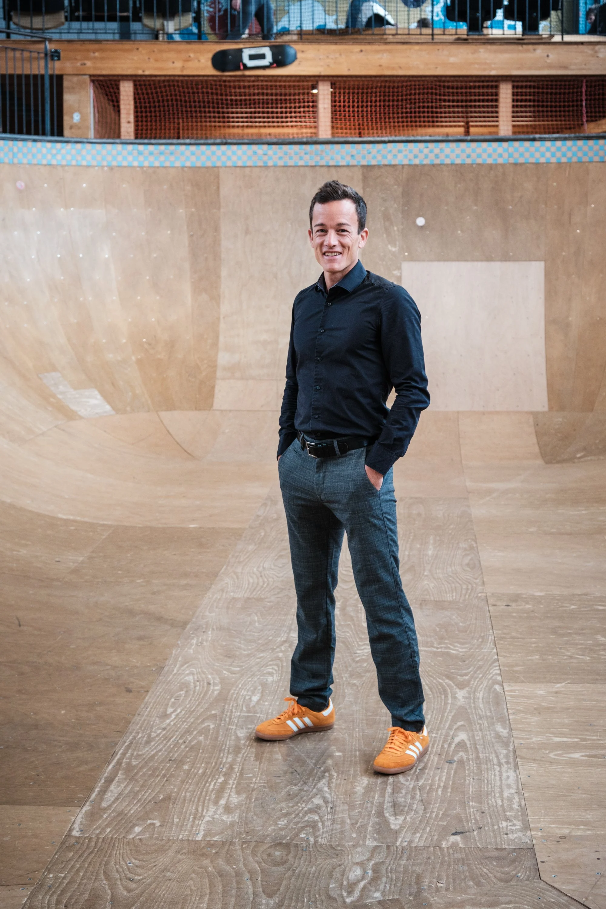 Junge Mann in schwarzem Hemd und grauen karierten Hosen mit orangen Turnschuhen, der auf einem Skateboard in einer Indoor-Skatehalle steht.