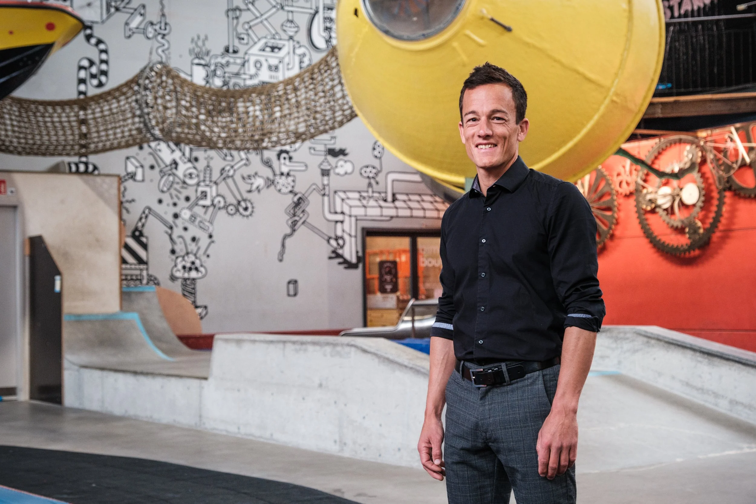 Ein Mann in dunklem Hemd und karierten Hosen steht in einem Indoor-Skatepark mit bunten Elementen und einer Wand mit Graffiti-Kunst im Hintergrund.