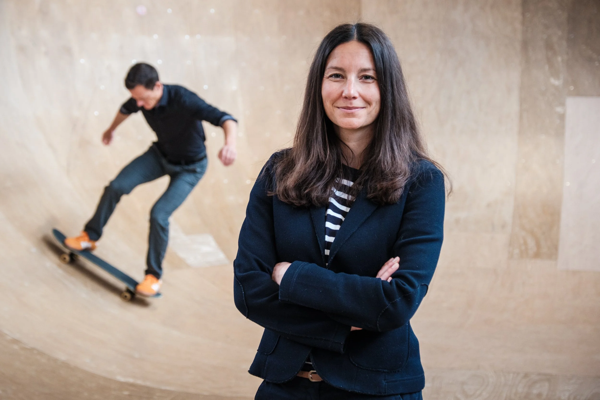 Eine Frau steht mit verschränkten Armen vor einer Skateboard-Rampe, im Hintergrund fährt eine junge Person Skateboard.