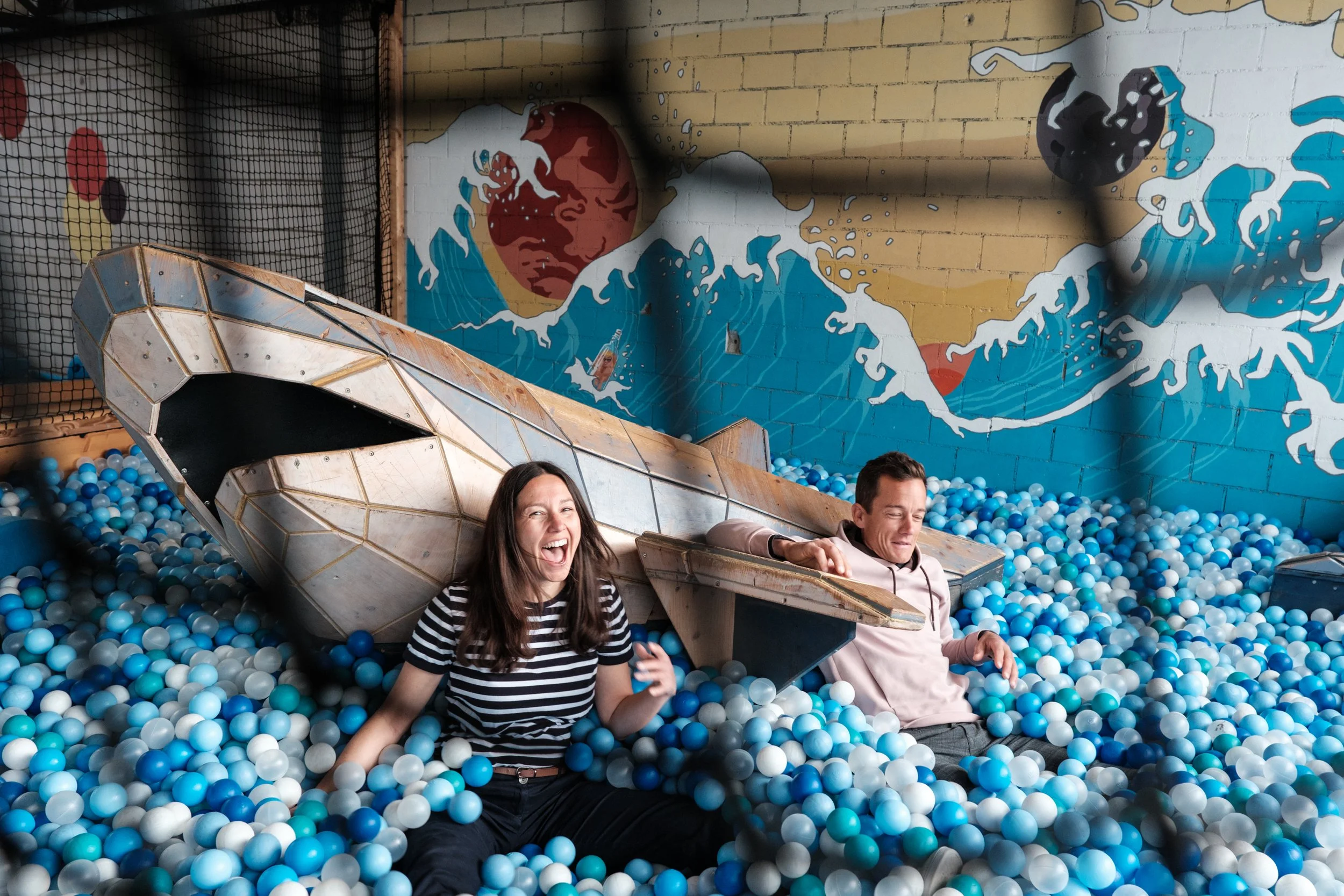 Zwei Menschen in einem **Bällebad** mit einem großen **Schiff-ähnlichen Spielzeug**. Im Hintergrund eine Wand mit **Streetart-Gemälde** eines großen **Wellen** und einer **kopflosen Figur**. Die Frau lächelt lachend, der Mann wirkt amüsiert.