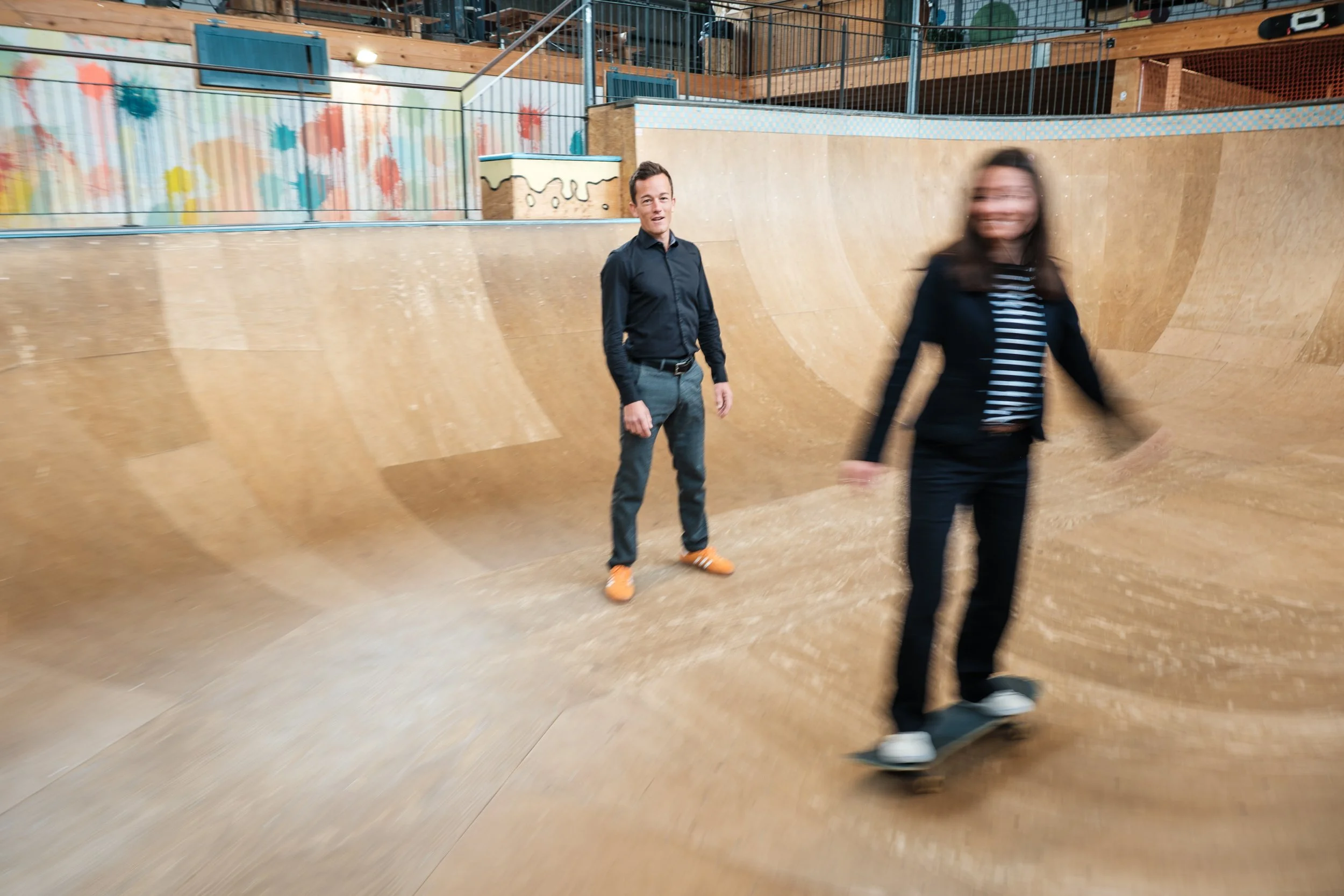 Zwei Menschen, ein Mann und eine Frau, in einer Indoor-Skatehalle, der Mann steht still und die Frau fährt auf einem Skateboard, die Frau wirkt im Vordergrund durch Bewegung unscharf.