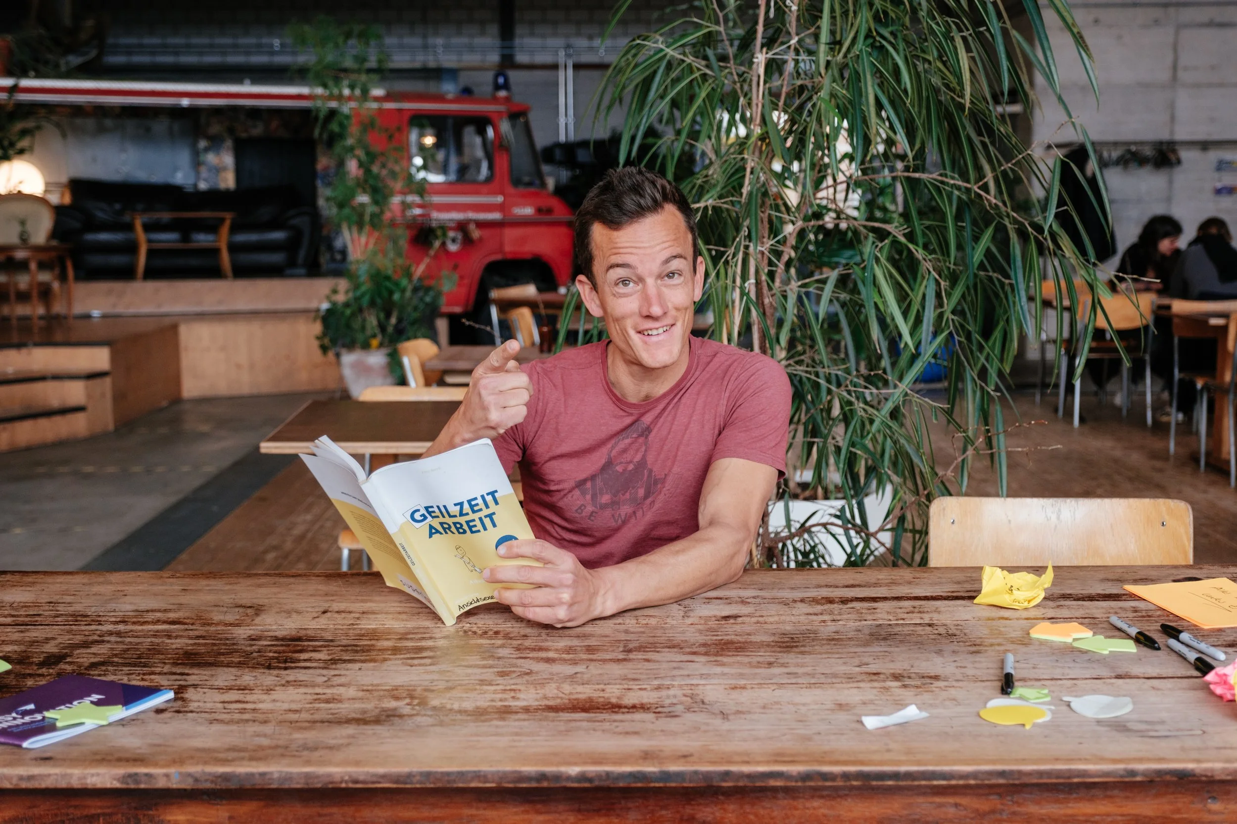 Mann sitzt an einem Holztisch in einem Café, hält ein Buch mit dem Titel 'GELZET ARBEIT', lächelt und zeigt auf das Buch, im Hintergrund Pflanzen und andere Gäste.