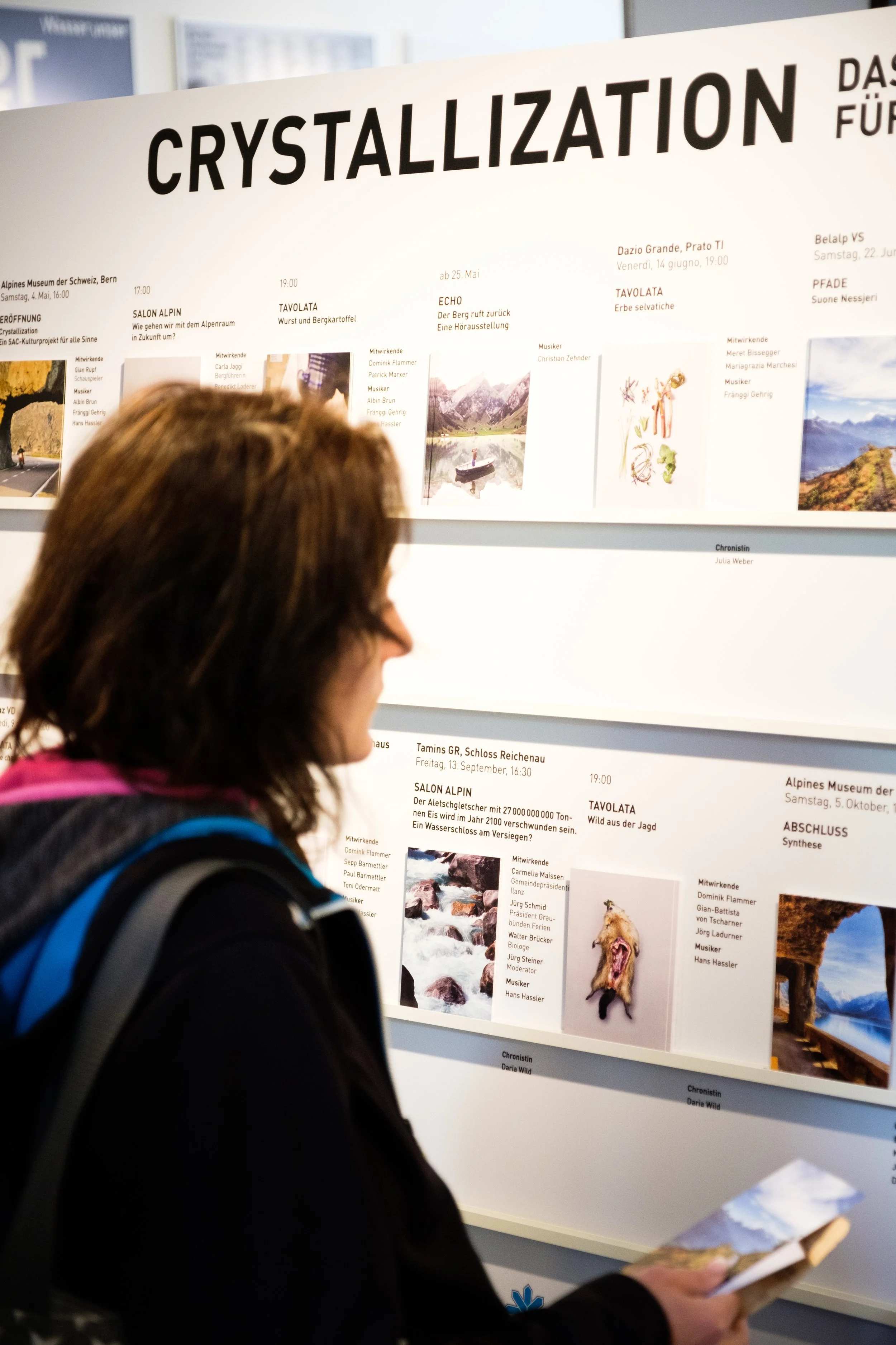 Eine Frau betrachtet ein Poster zur Ausstellungsreihe 'CRYSTALLIZATION' am Alpinmuseum in der Schweiz, Bern, mit weiteren Informationen über Veranstaltungen und Mitwirkende.